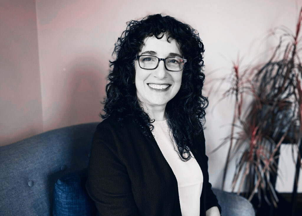 A photo of Allison Plesur - A woman with curly hair and glasses wearing a cardigan and sitting in a chair.